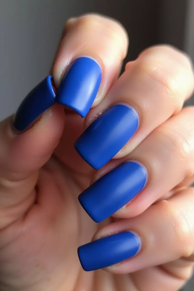 Matte Blue Finish Close-up of a woman's hand with five fingers showing cobalt blue nails with completely matte finish, no shine, creating a velvety texture appearance in natural light.