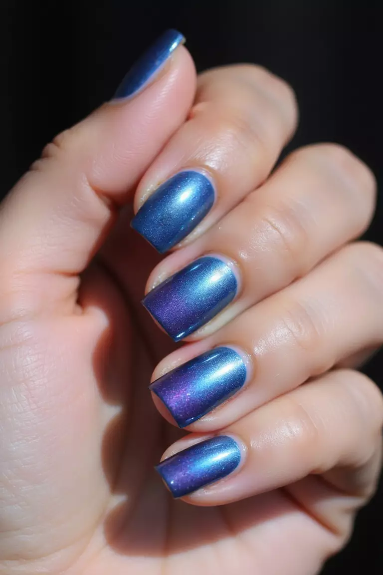 Holographic Blue Close-up of a woman's hand with five fingers displaying iridescent blue nail polish that shifts colors from blue to purple when moved in light, with a metallic rainbow effect.