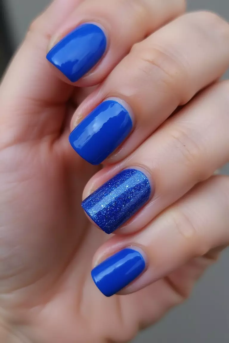 Glitter Blue Accent Nails Close-up of a woman's hand with five fingers featuring solid cobalt blue nails with ring and thumb fingers covered in matching blue glitter polish that sparkles in the light.