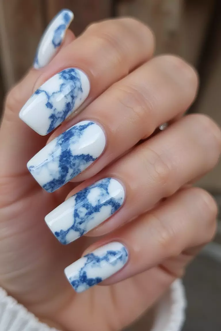 Blue Marble Effect Close-up of a woman's hand with five fingers showing white nails with swirling blue marble patterns resembling natural stone, with glossy topcoat reflecting light.