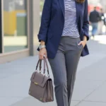 A beautiful woman in a navy blazer, striped long-sleeve top, tailored gray trousers, and brown loafers, she carries a structured handbag.
