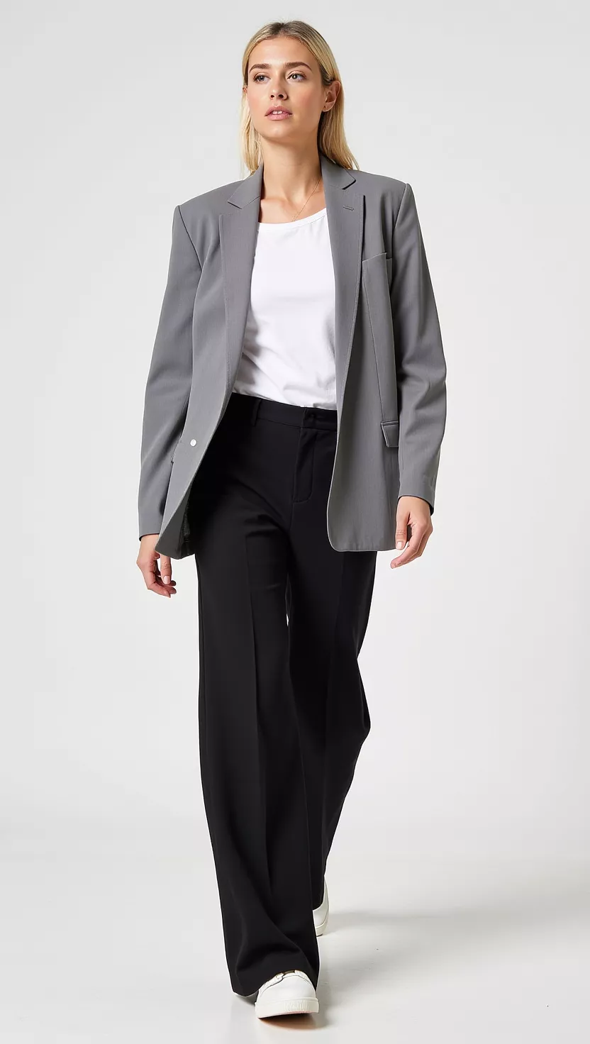 A beautiful woman in an oversized grey blazer, white t-shirt, black wide-leg trousers, and white sneakers, she is walking confidently.