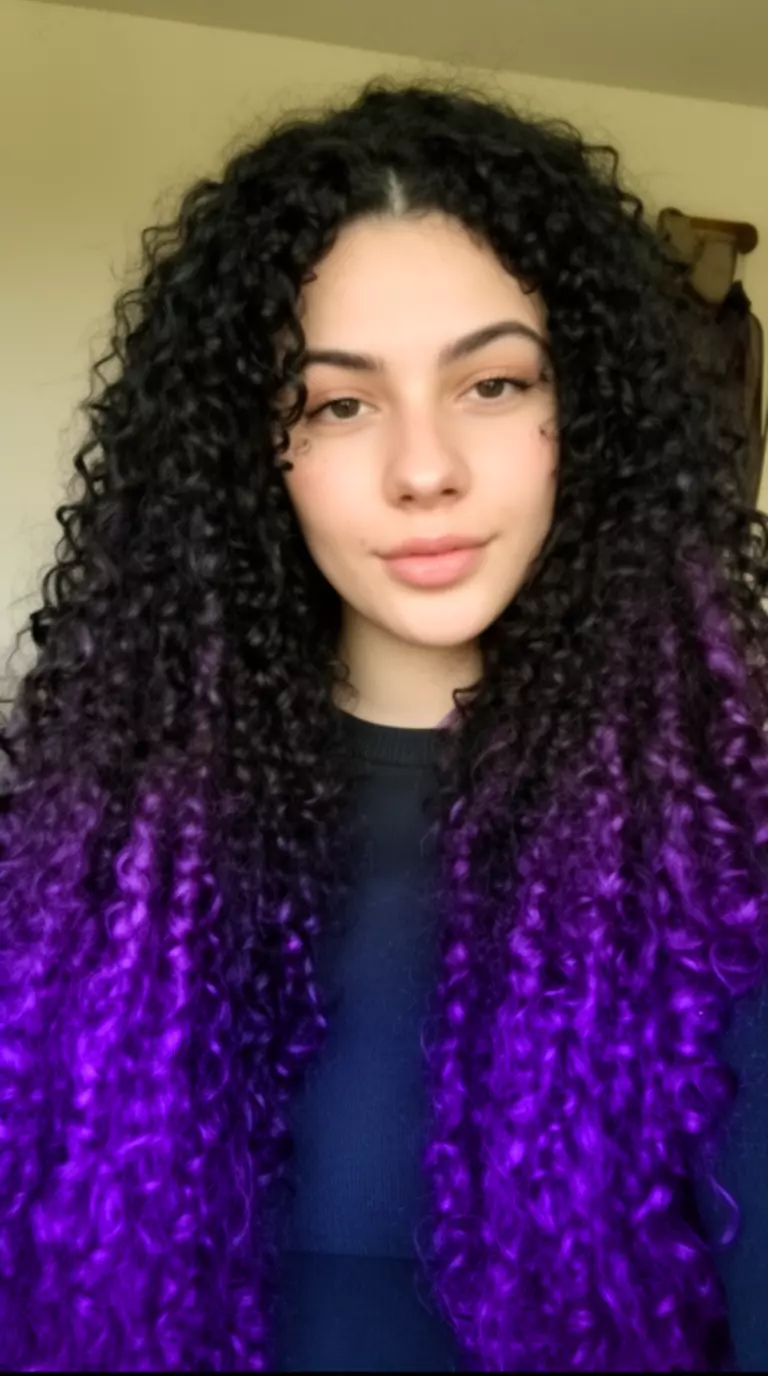 selfie of a 26-year-old woman wearing a dark top with a black curly hair with purple streaks colored very long, voluminous hairstyle, front-facing view, minimal background