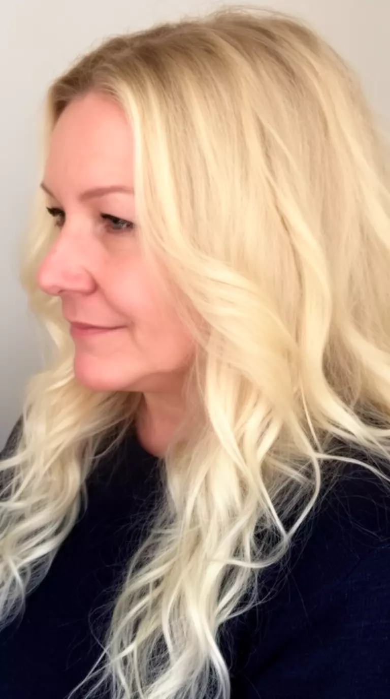 casual close-up photo of a 40-year-old woman wearing a dark top with a bright blonde colored long, wavy hairstyle, profile view, minimal background
