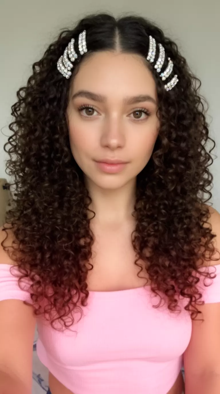 selfie of a 27-year-old woman wearing a light pink off-the-shoulder top with voluminous, dark brown curly hair and multiple rhinestone clips, minimal background