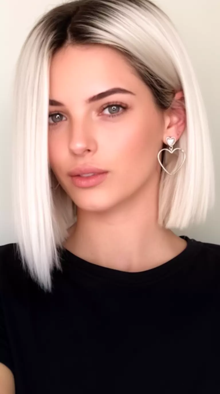 casual close-up photo of a 29-year-old woman wearing a black top and heart earrings with a platinum blonde straight sharp bob haircut featuring dark roots, minimal background