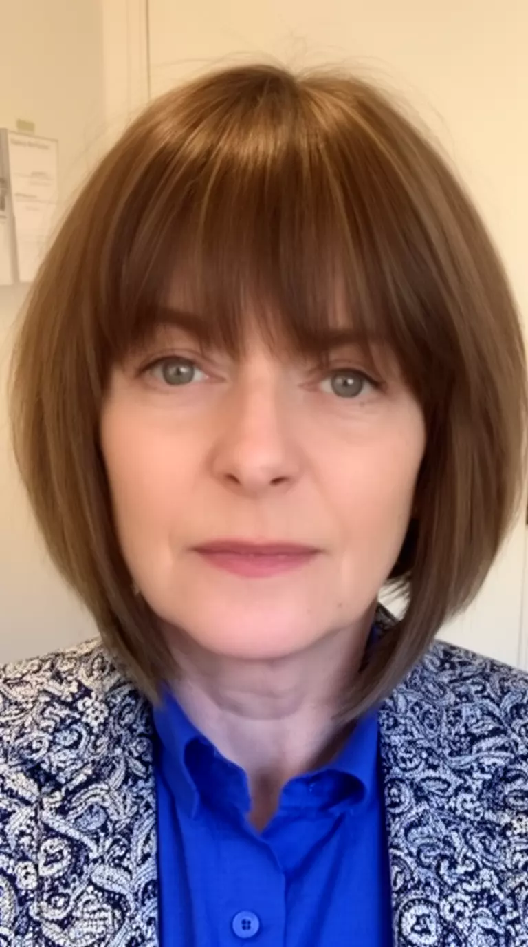 casual close-up photo of a 42-year-old woman wearing a blue shirt and patterned jacket with a layered medium-length bob featuring long sweeping bangs, minimal background