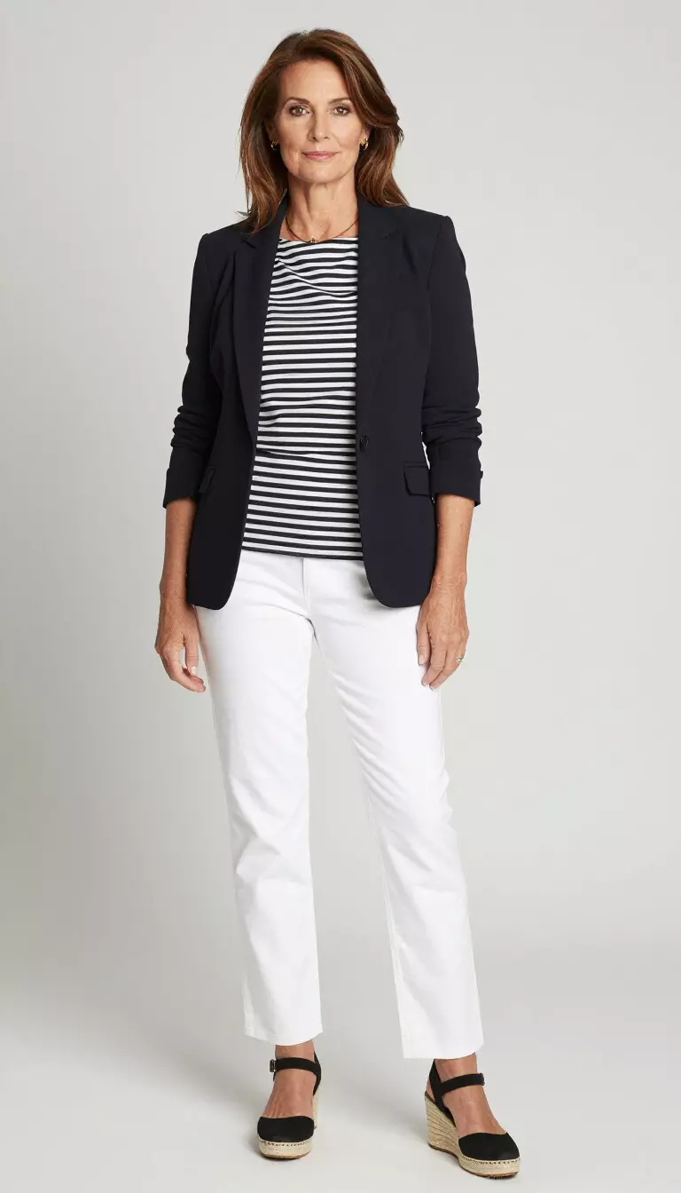 A beautiful 62-year-old woman in a black and white horizontally striped top, a black blazer, white pants, and black wedge espadrilles.