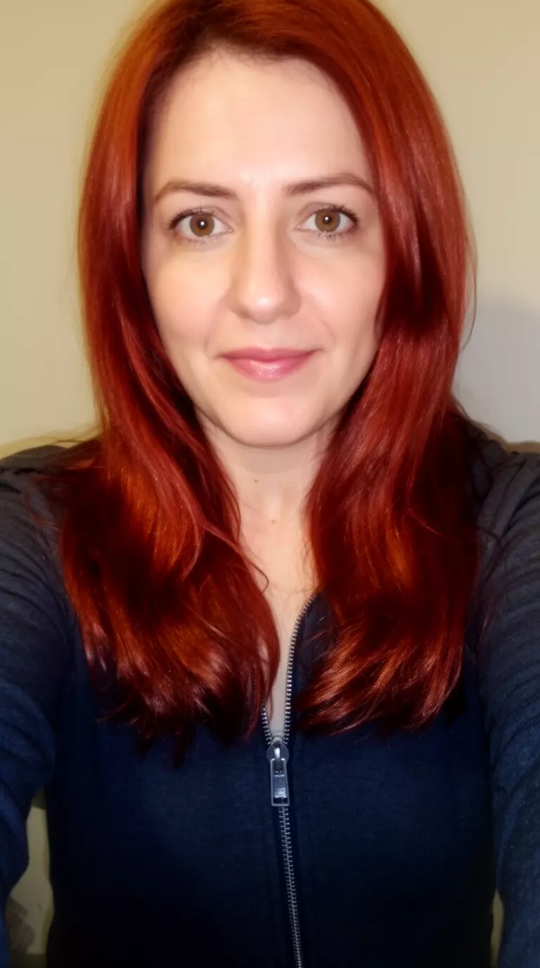 selfie of a 35-year-old woman wearing a dark zip-up top with a glossy deep copper red colored medium-long hairstyle, front-facing view, minimal background