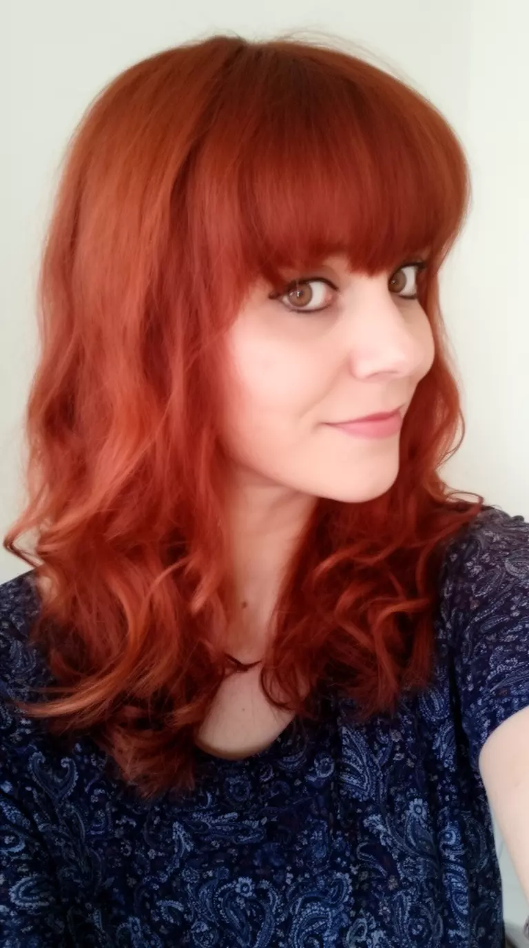 selfie of a 33-year-old woman wearing a patterned dark top with a vibrant reddish-brown colored medium-long wavy hairstyle with bangs, side angled view, minimal background