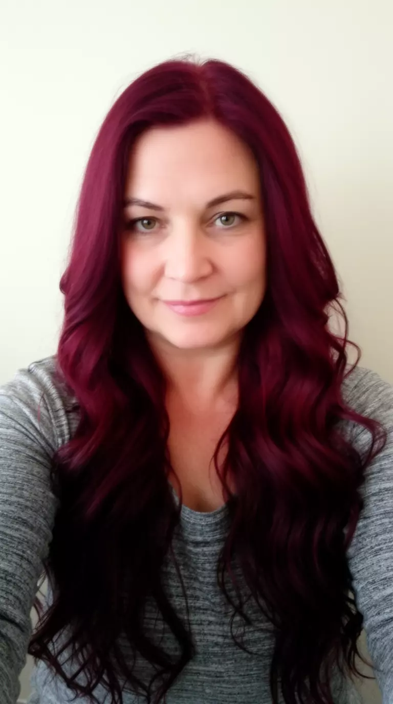 selfie of a 38-year-old woman wearing a gray top with a burgundy colored long wavy hairstyle, front view, minimal background