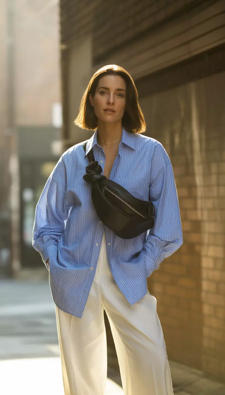 A realistic photo of a woman dressed in an oversized light blue and white pinstripe button down shirt worn open over white wide leg trousers with a knotted black bag.