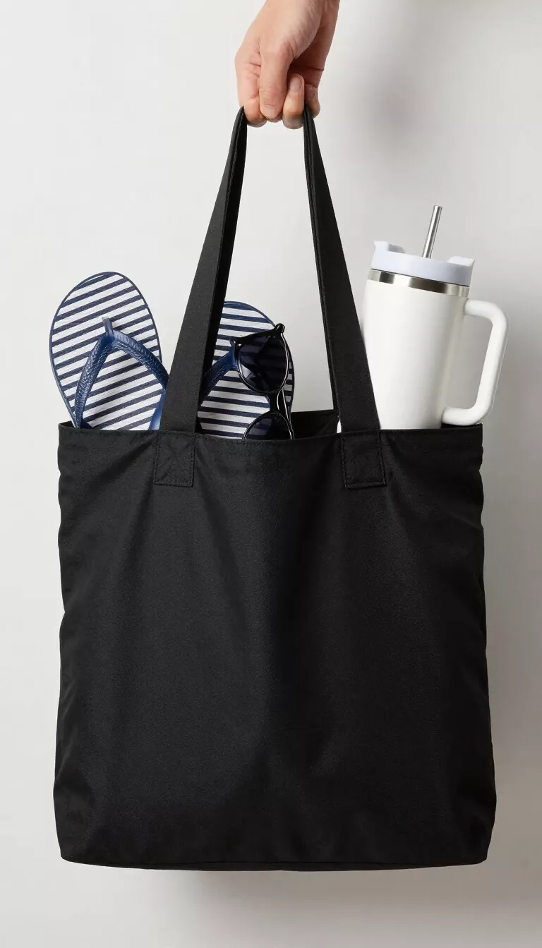 A solid black functional fabric tote bag holding a pair of striped flip flops, sunglasses, and a large white insulated tumbler with a handle sticking out of the top.