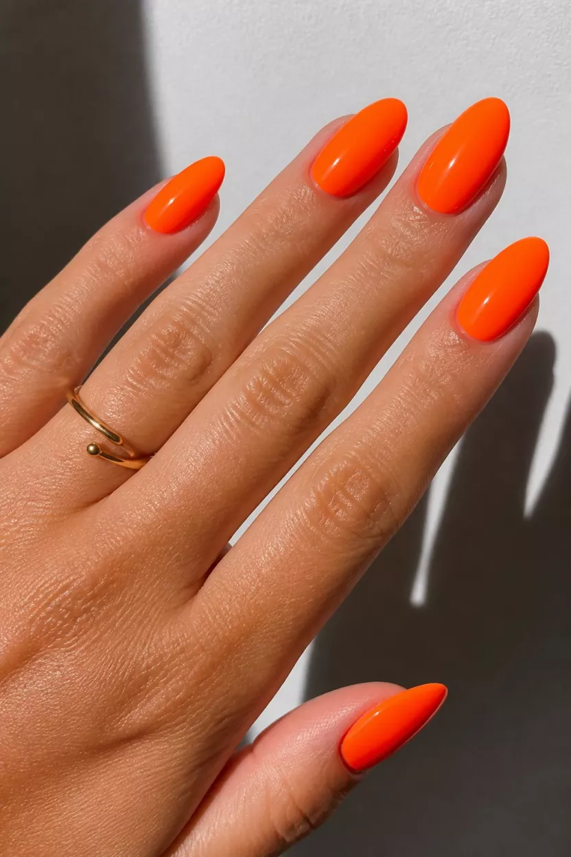 Neon Orange Glow close-up shot of a woman’s hand with five fingers showing nails with almond-shaped nails painted a striking, glossy neon orange highlighted by dramatic shadows and a simple gold ring