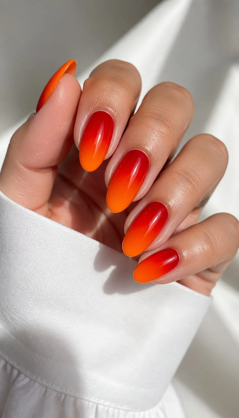 close-up shot of a woman’s hand with five fingers showing long oval-shaped nails with a striking ombre effect, transitioning from deep red at the base to bright orange at the tips, woman wearing a white garment.
