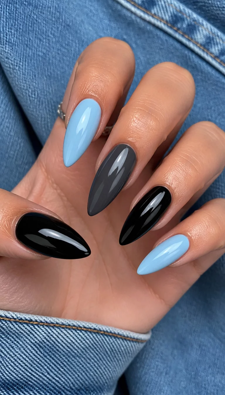 Denim Blue And High Shine close-up shot of a woman’s hand with five fingers showing nails with a mix of light blue almond nails, high shine stiletto charcoal nails, and glossy black nails, denim background.