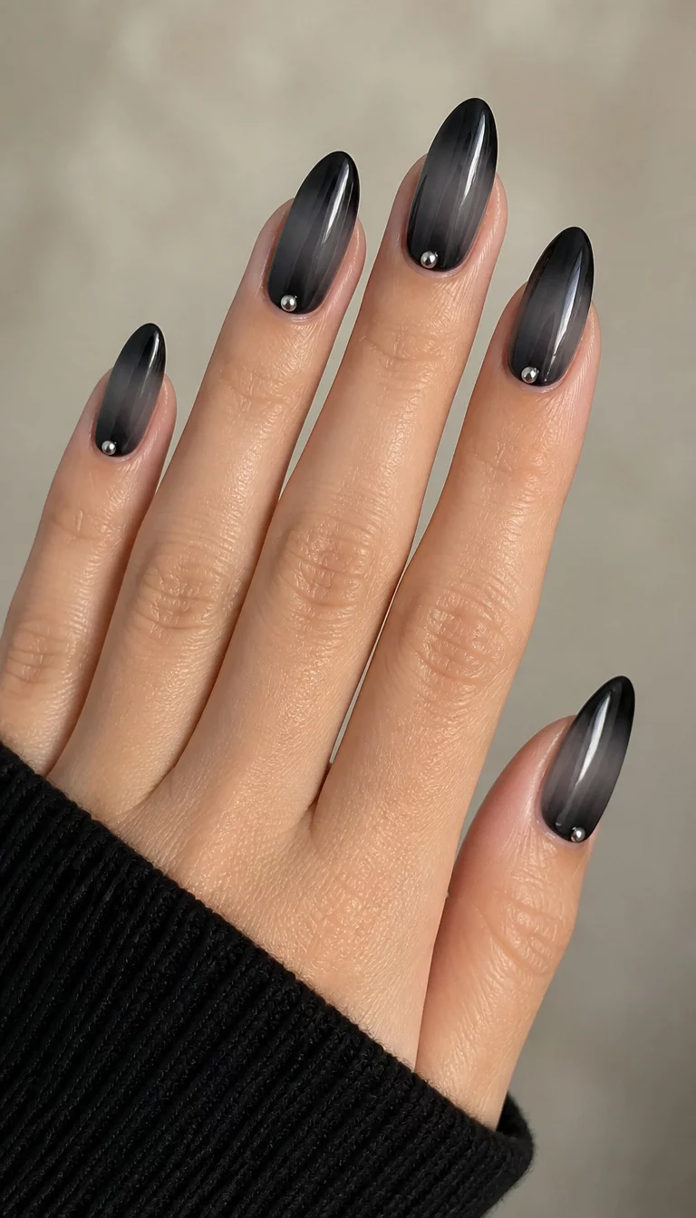 Ombre Studded Charcoal close-up shot of a woman’s hand with five fingers showing nails with almond-shaped nails with a dark grey to black ombre effect and small silver studs at the cuticle, soft neutral background.