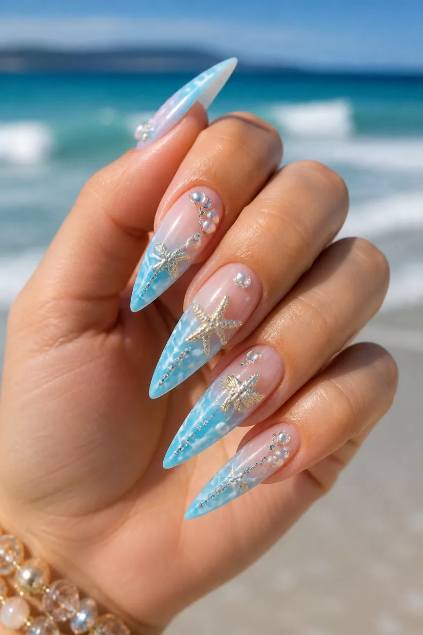 close-up shot of a woman’s hand with five fingers showing nails with long stiletto shaped nails featuring a light blue ombre with white wave accents adorned with pearls and rhinestones and starfish, beach ocean theme background.