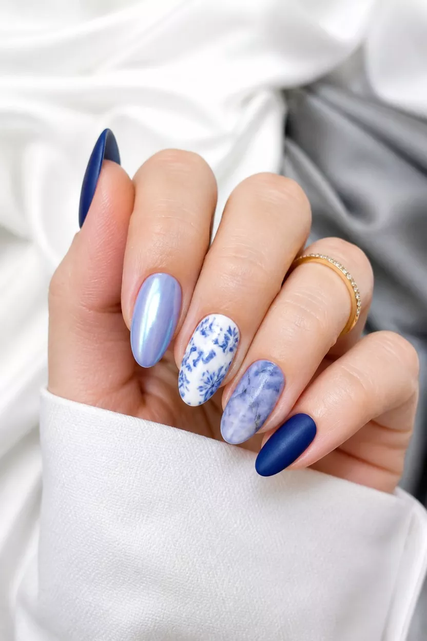 close-up shot of a woman’s hand with five fingers showing nails with varied blue nail designs including pearlescent blue and blue floral transfers and blue marble and deep matte blue, white and gray fabric background.