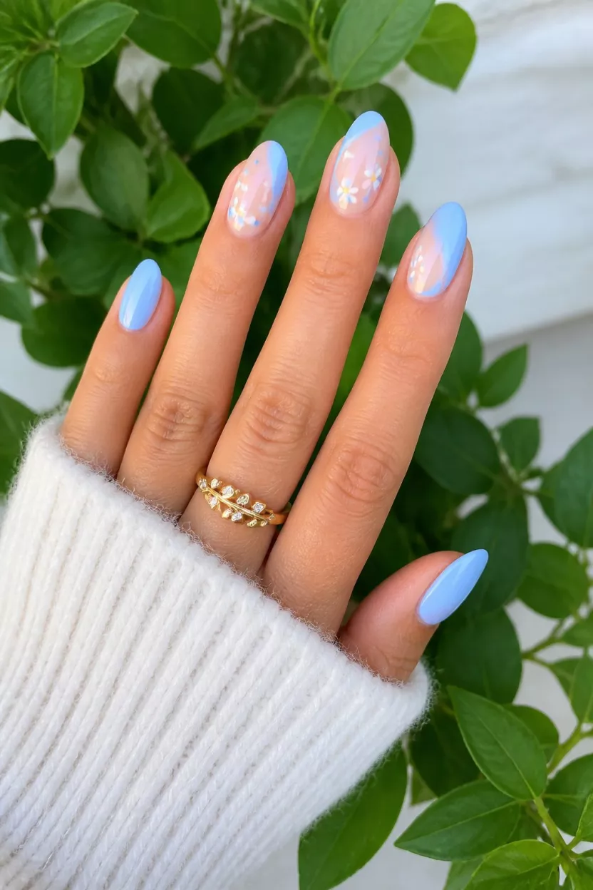 close-up shot of a woman’s hand with five fingers showing nails with almond shaped nails in pastel blue with white daisy accents on a nude base, green foliage and white sweater background.