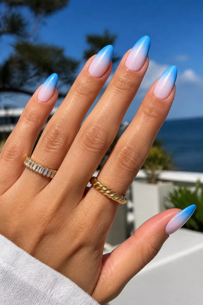 close-up shot of a woman’s hand with five fingers showing nails with almond shaped natural nails showing a vibrant blue to white ombre effect, bright outdoor setting with gold rings.