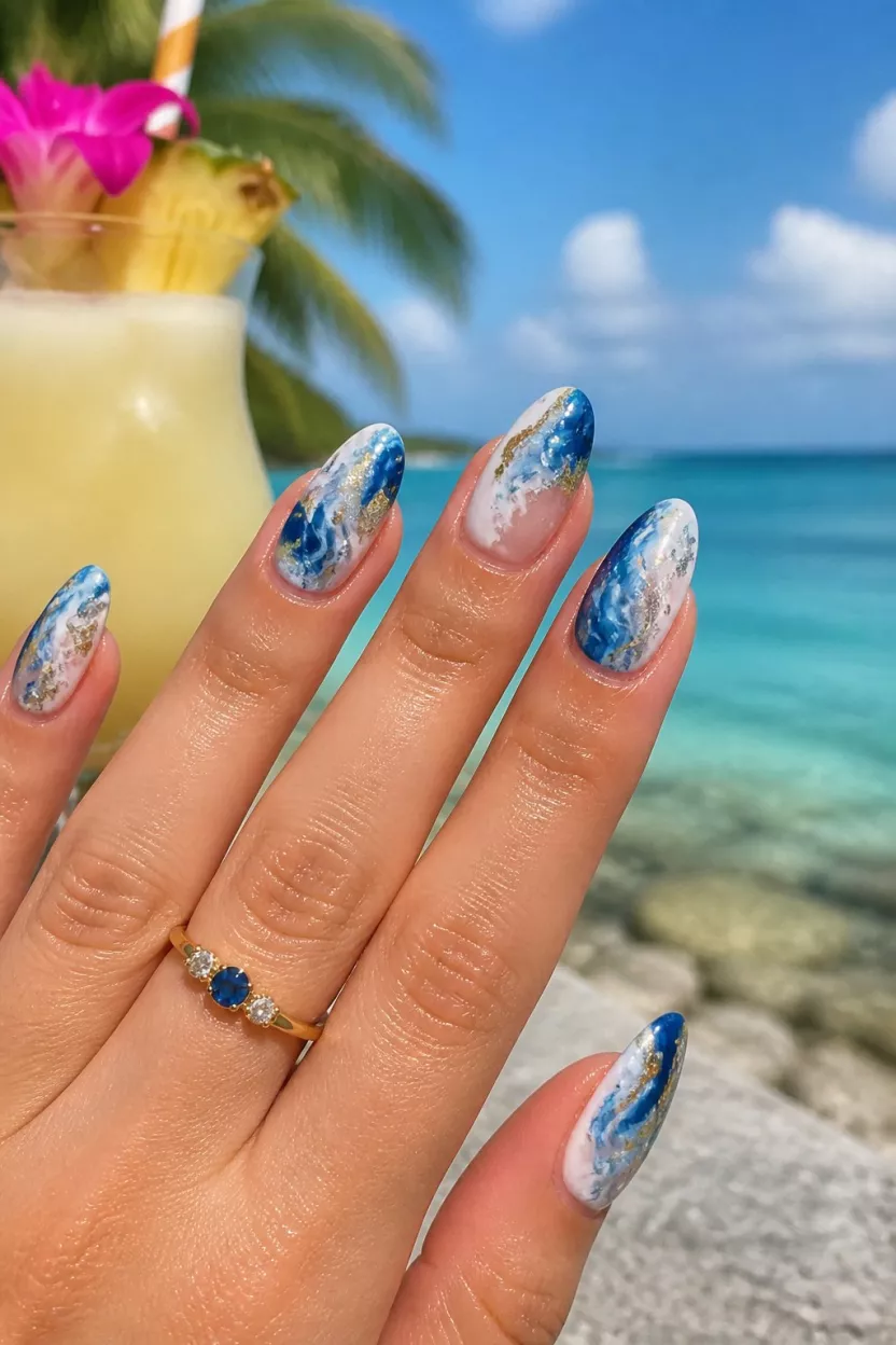 close-up shot of a woman’s hand with five fingers showing nails with almond shaped nails with blue and white wave designs and silver and gold foil accents, tropical beach backdrop with a cocktail.