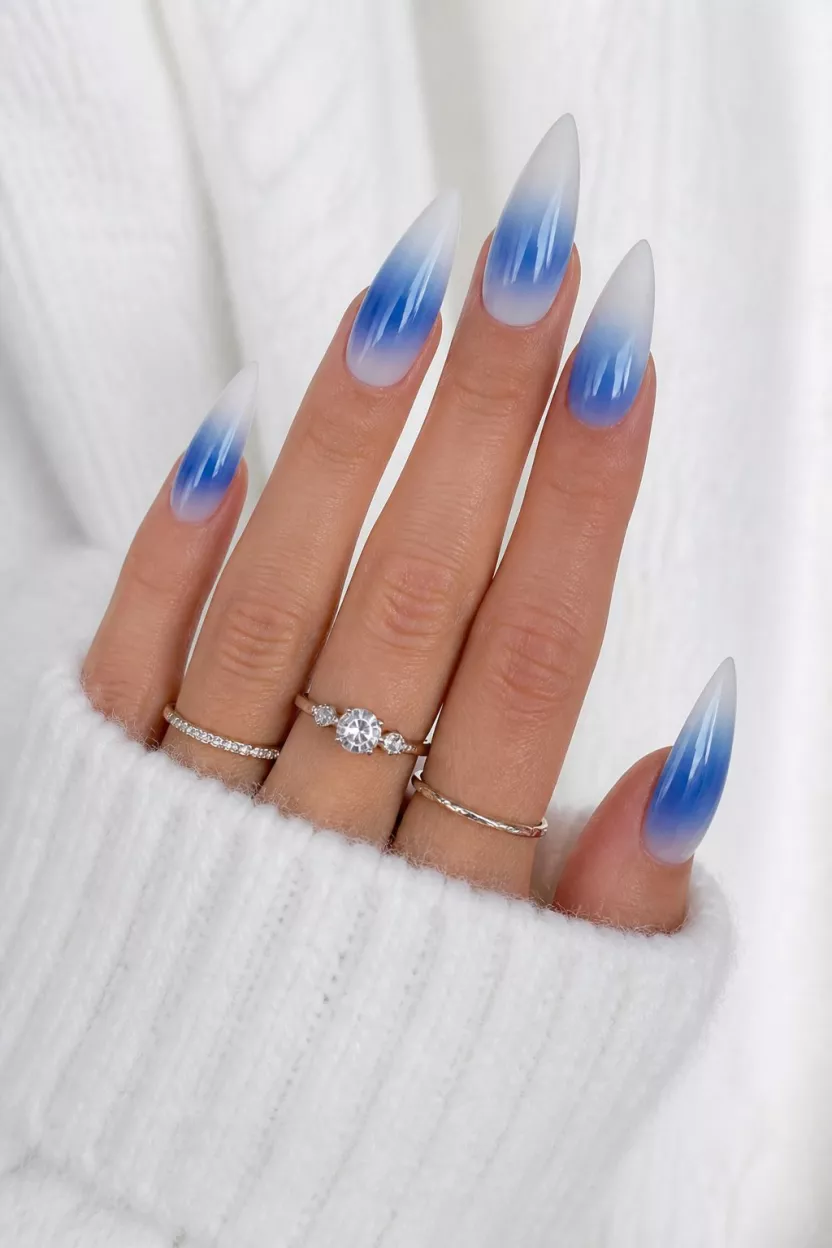 close-up shot of a woman’s hand with five fingers showing nails with stiletto nails exhibiting a striking blue and white blur or ombre effect, white sweater background with silver rings.