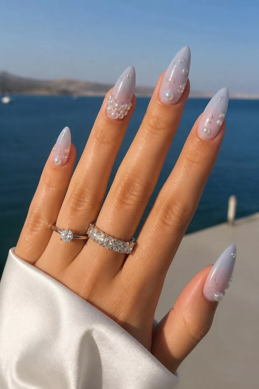 close-up shot of a woman’s hand with five fingers showing long, pointed nails in a milky, bluish-white shade with ornate 3D pearl and crystal details, held outdoors near water.