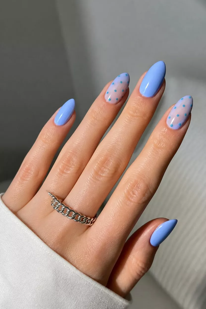 close-up shot of a woman’s hand with five fingers showing almond-shaped nails in light periwinkle blue, with some featuring sheer bases detailed with small teal polka dots, worn with a silver ring.