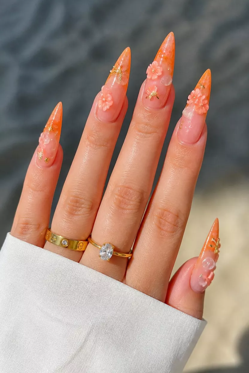 close-up shot of a woman’s hand with five fingers showing long, pointed nails with a bright orange ombré fade at the tips over a sheer base, featuring 3D flower and starfish sea charms.