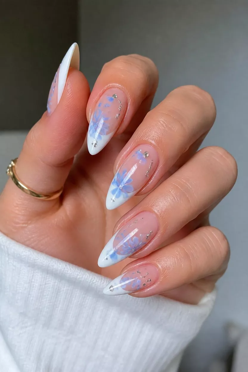 close-up shot of a woman’s hand with five fingers showing long, pointed almond nails with sharp white French tips accented by delicate, hand-painted light blue flowers and small rhinestones.