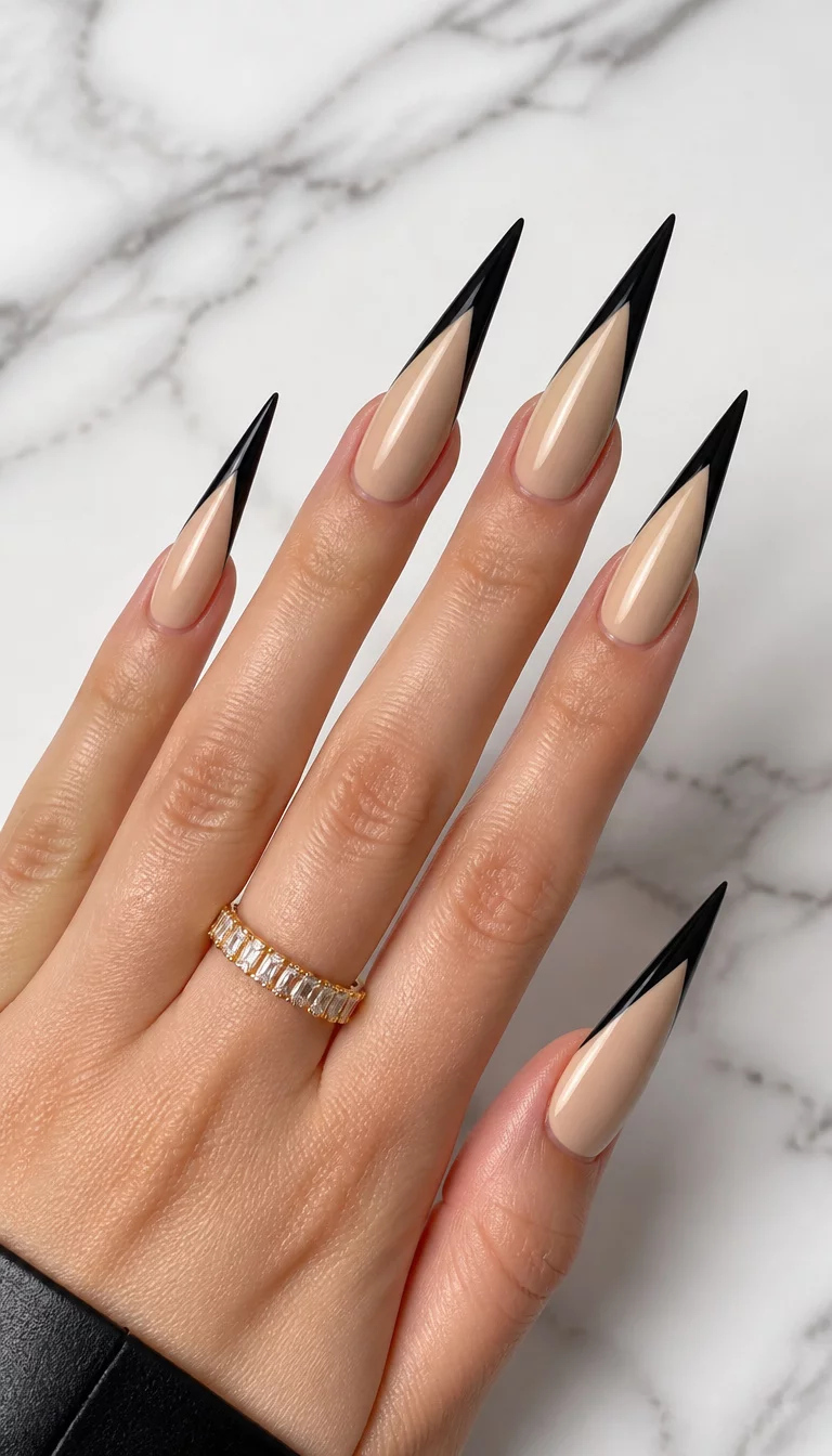 close-up shot of a woman’s hand with five fingers showing extra-long stiletto nails with a neutral beige base and a sharp black V-shaped French tip design, set against a white marble background.