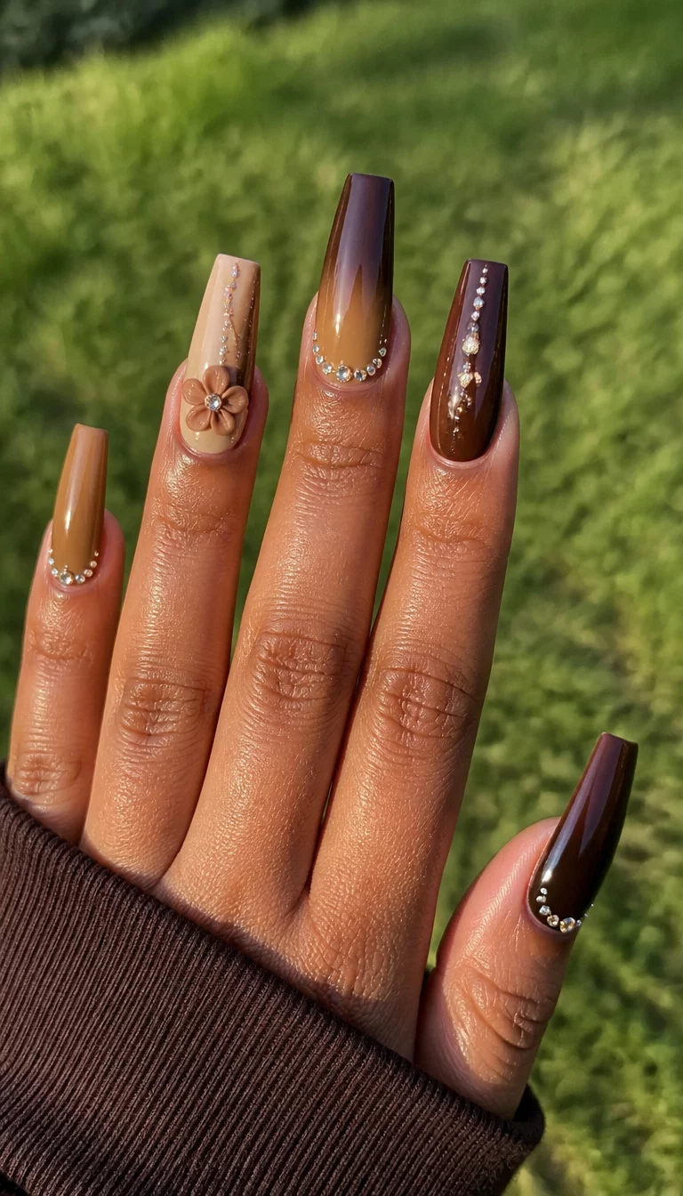 Brown Gradients And 3D Florals close-up shot of a woman’s hand with five fingers showing long coffin nails with a gradient of chocolate brown and tan tones, rhinestones, and a 3D flower design, outdoor grassy area background.