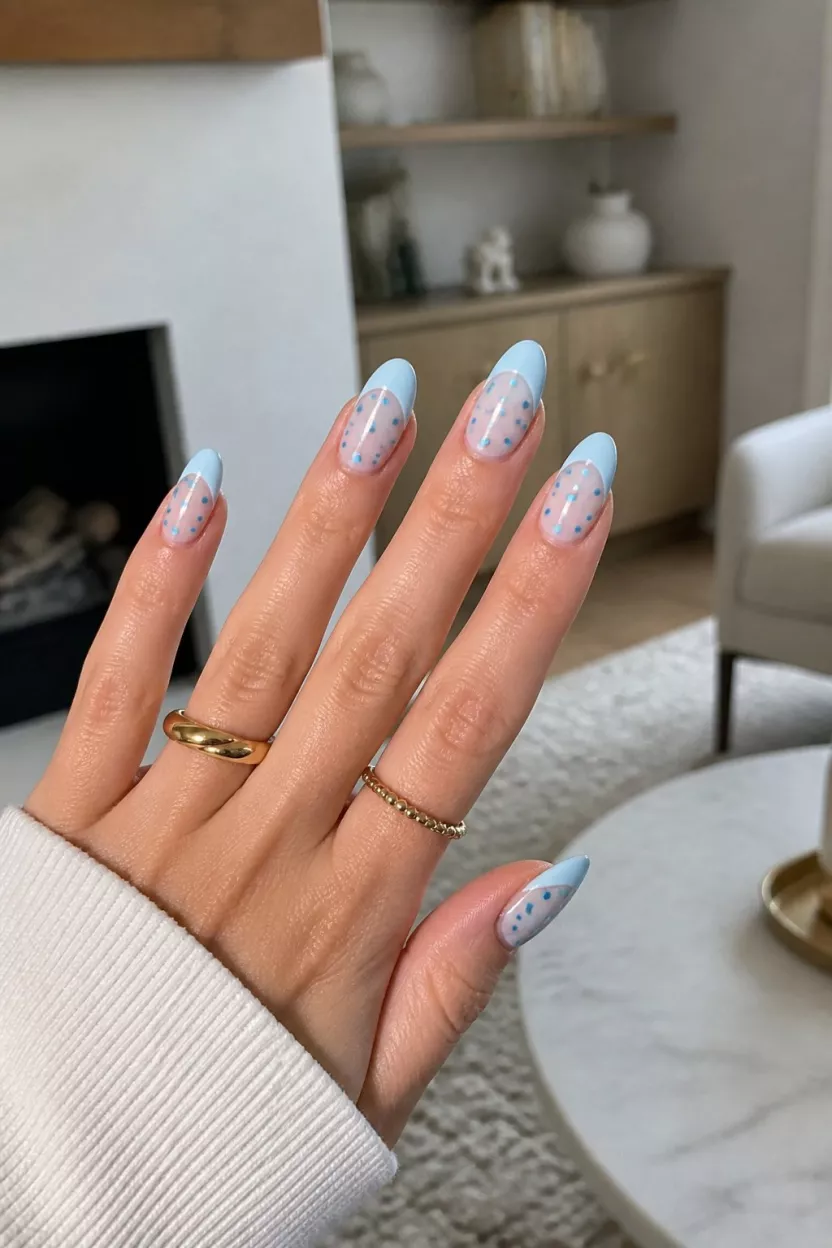 close-up shot of a woman’s hand with five fingers showing almond-shaped nails in soft pale blue with sheer bases and tiny teal polka dots, simple jewelry, living room background.