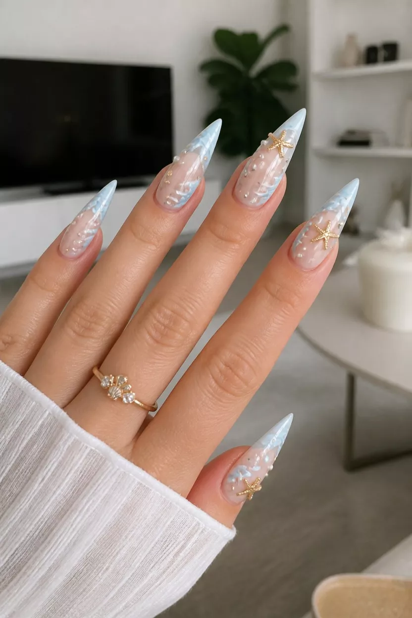 close-up shot of a woman’s hand with five fingers showing stiletto nails with a sheer base, light blue wave designs, white pearls, and gold starfish charms, living room background.