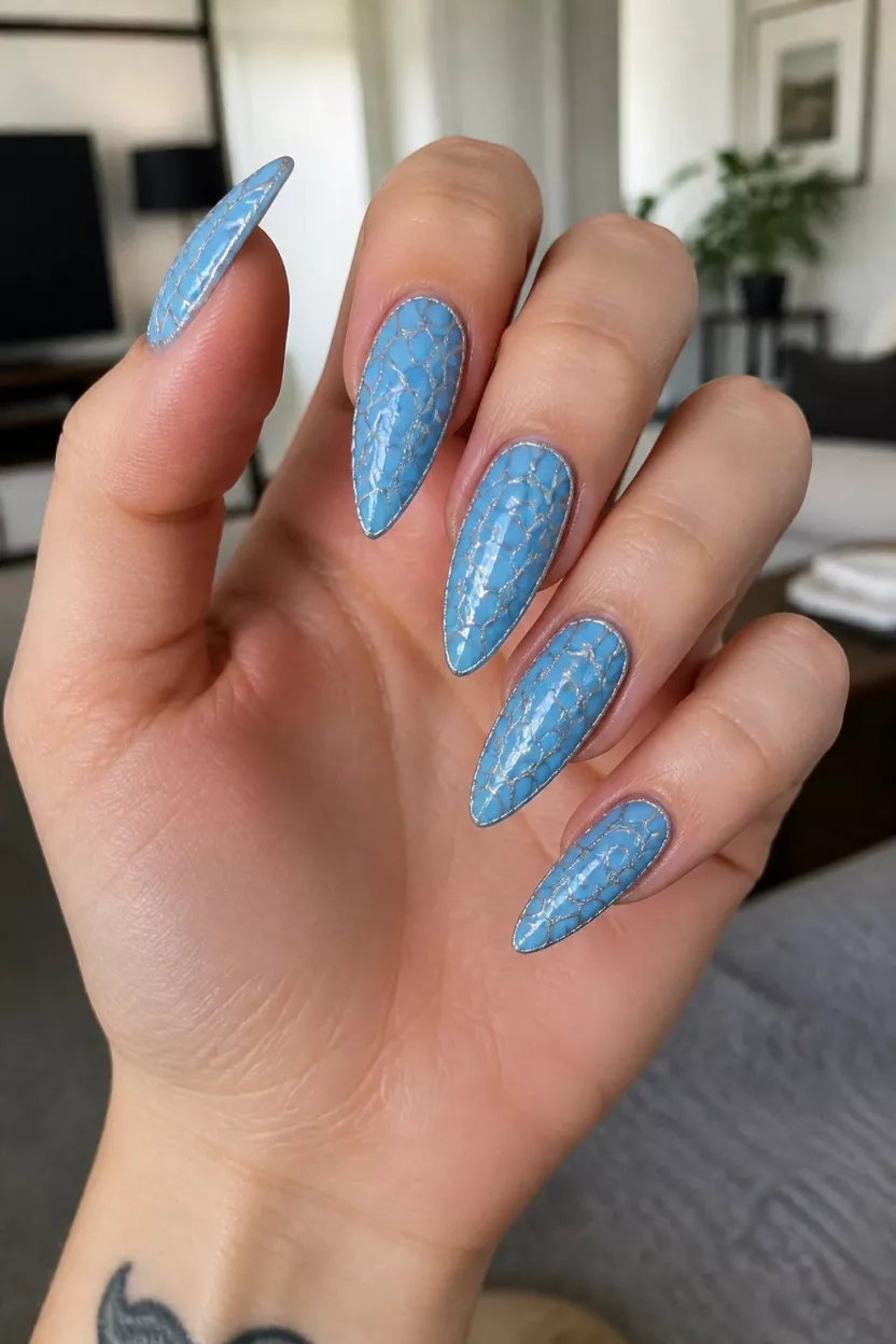 close-up shot of a woman’s hand with five fingers showing light blue stiletto nails with a textured scaly design outlined in silver, living room background.