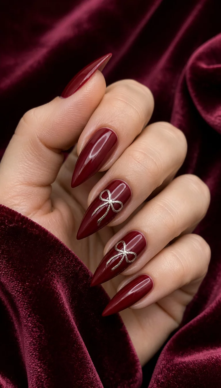 close-up shot of a woman’s hand with five fingers showing stiletto nails in a deep red glossy polish, with two accent nails featuring delicate silver bow designs, soft velvet fabric background.