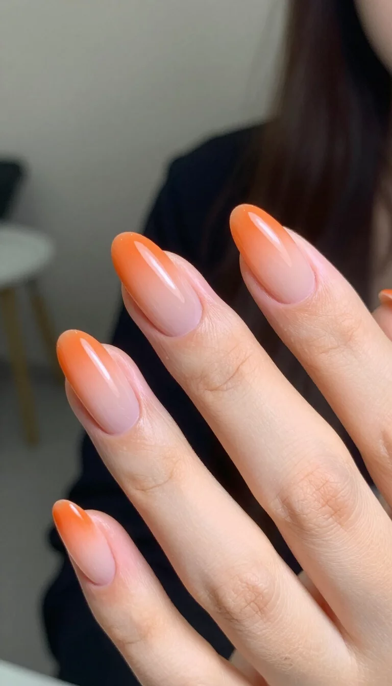 close-up shot of a woman’s hand with five fingers showing nails with almond-shaped nails with an orange ombre fade transitioning to a sheer nude, blurred indoor setting.