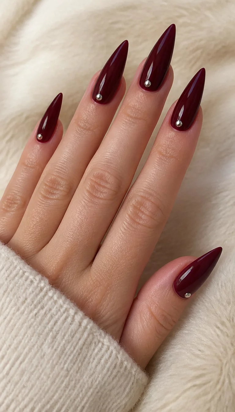 Studded Burgundy Stiletto close-up shot of a woman’s hand with five fingers showing stiletto-shaped, deep burgundy nails with individual silver studs at the base of each nail, soft light-colored surface.