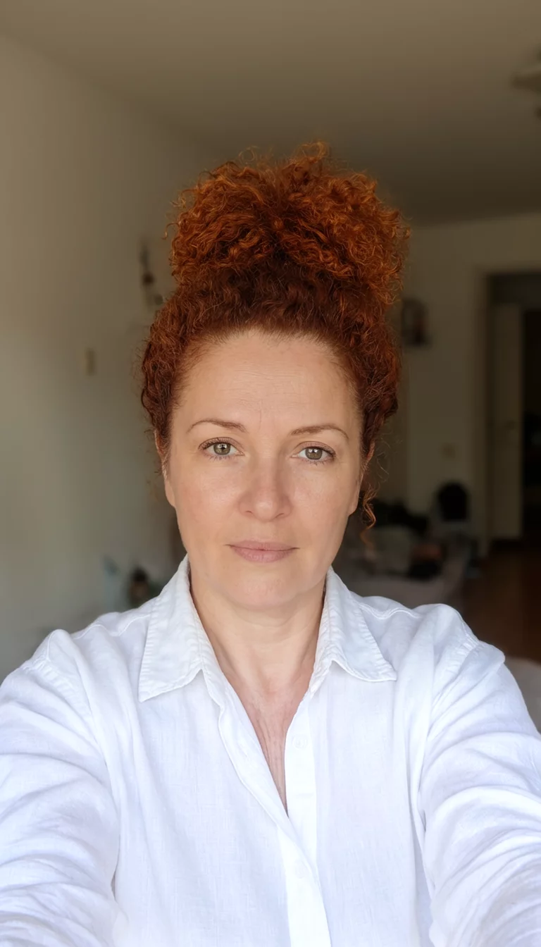 selfie of a 41-year-old woman wearing a white button-down shirt with a reddish-brown colored curly hair in a high bun hairstyle, front view, indoor setting