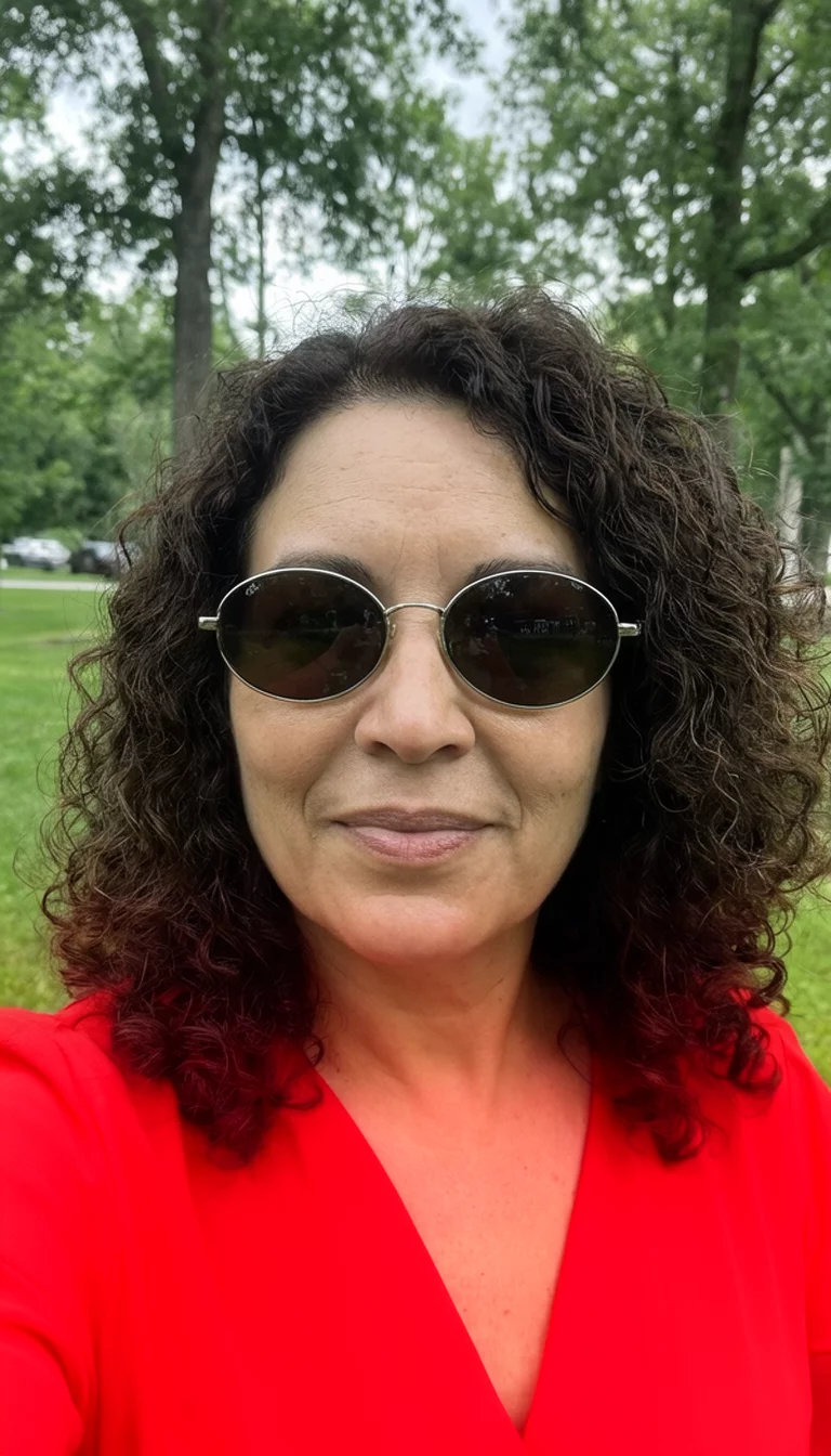 selfie of a 35-year-old woman wearing a bright red top and oval sunglasses with a dark brown colored curly hairstyle, front view, outdoor setting