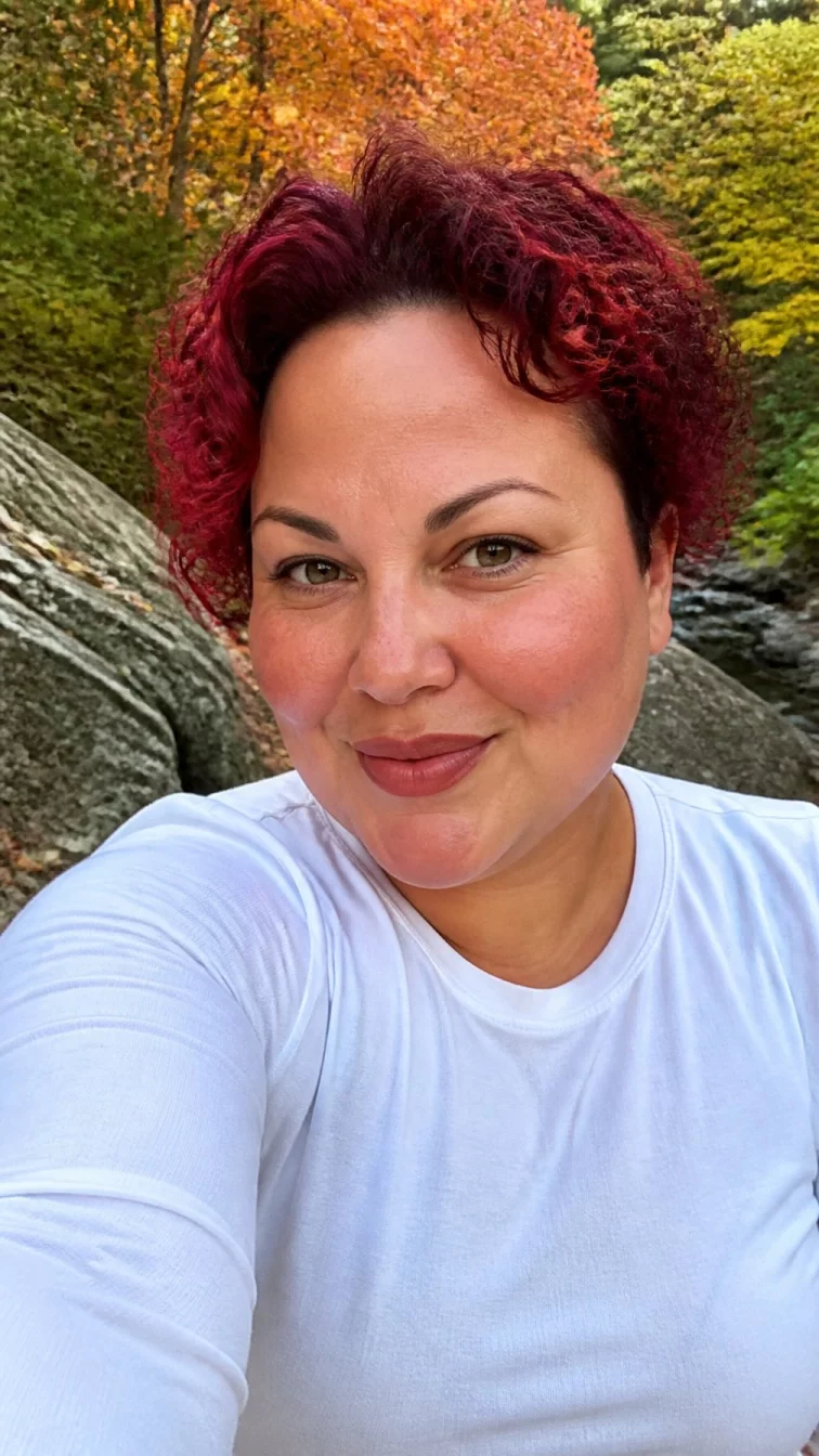 selfie of a 44-year-old Latina plus size woman with a chubby face, tan, aquiline nose, thin arched eyebrows, and wide lips. Her face features widow's peak hairline, slightly flushed cheeks, and subtle natural facial asymmetry. She has almond-shaped green eyes, rounded cheeks, closed-mouth smile, playful. She is athletic and is wearing a white long sleeve t-shirt with ruby grapefruit pinkish-red short curly hair, warm indoor lighting, outdoor fall background