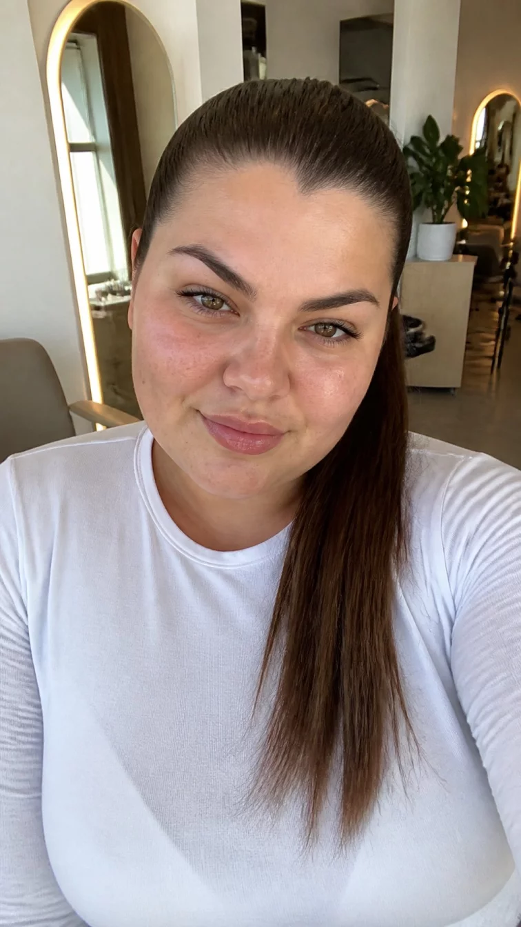 selfie of a 23-year-old Southern European plus size woman with a chubby face, olive, upturned nose, thin arched eyebrows, and heart-shaped lips. Her face features widow's peak hairline, subtle acne marks, and subtle natural facial asymmetry. She has almond-shaped amber eyes, high cheekbones, neutral expression, cheerful. She is curvy and is wearing a white long sleeve t-shirt with iced mocha latte cool brunette sleek high ponytail hair, soft window light, hair salon