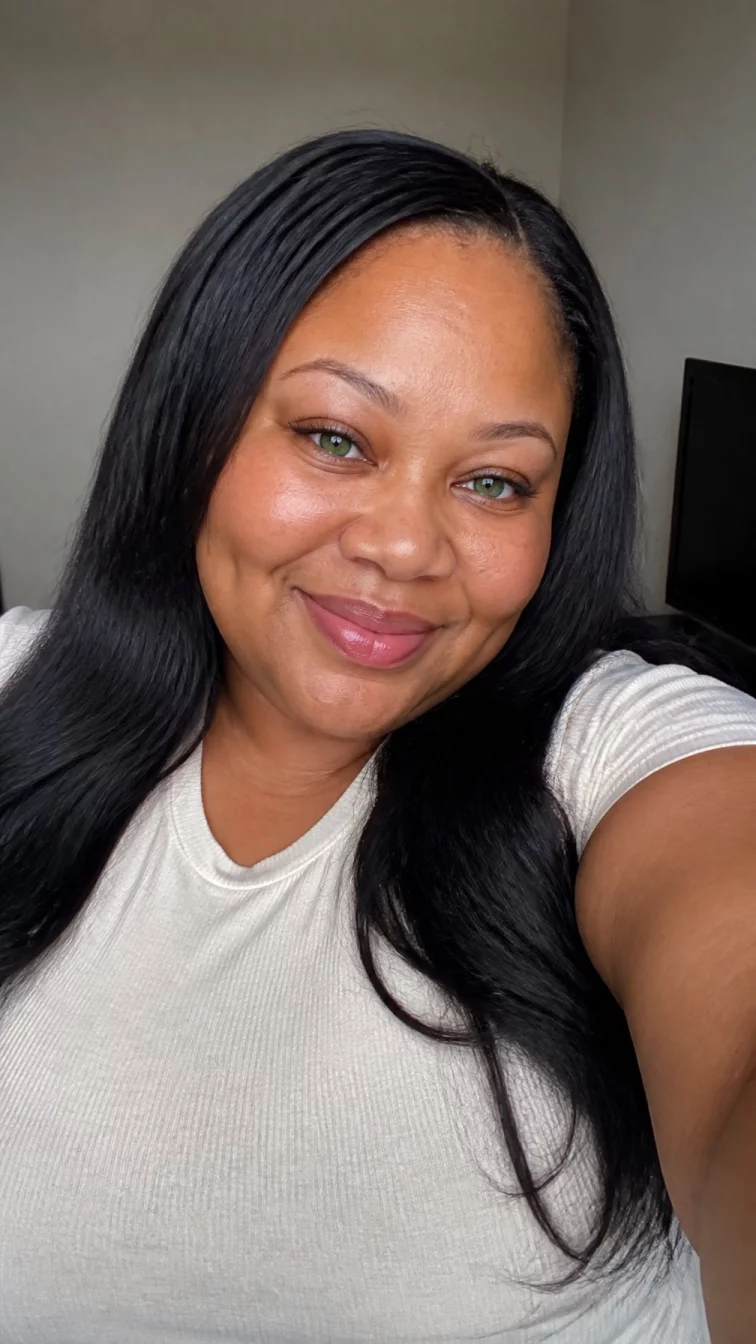 selfie of a 45-year-old Black African plus size woman with a chubby face, light, narrow nose, feathered eyebrows, and heart-shaped lips. Her face features dimples, subtle acne marks, and subtle natural facial asymmetry. She has hooded green eyes, soft cheekbones, closed-mouth smile, cheerful. She is athletic and is wearing a cream basic crew neck t-shirt with midnight navy almost black blue sleek straight hair, soft window light, indoor