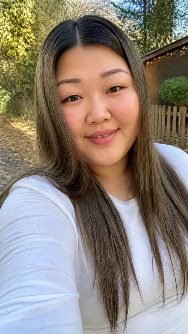 selfie of a 35-year-old East Asian plus size woman with a chubby face, dark brown, rounded nose, thin arched eyebrows, and heart-shaped lips. Her face features gap between front teeth, slightly flushed cheeks, and subtle natural facial asymmetry. She has hooded brown eyes, rounded cheeks, closed-mouth smile, relaxed. She is plus-size and is wearing a white long sleeve t-shirt with olive oil bronde muted green-brown long straight hair, soft window light, outdoor fall background