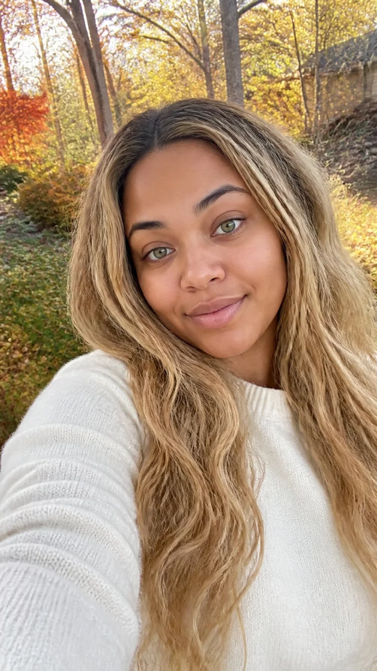 selfie of a 24-year-old Afro-Caribbean woman with a round face, tan, rounded nose, straight eyebrows, and full lips. Her face features slightly crooked smile, faint under-eye shadows, and subtle natural facial asymmetry. She has hooded green eyes, rounded cheeks, subtle smile, relaxed. She is curvy and is wearing a white cashmere sweater with creamy oat blonde long textured wavy hair, bright natural daylight, outdoor fall background