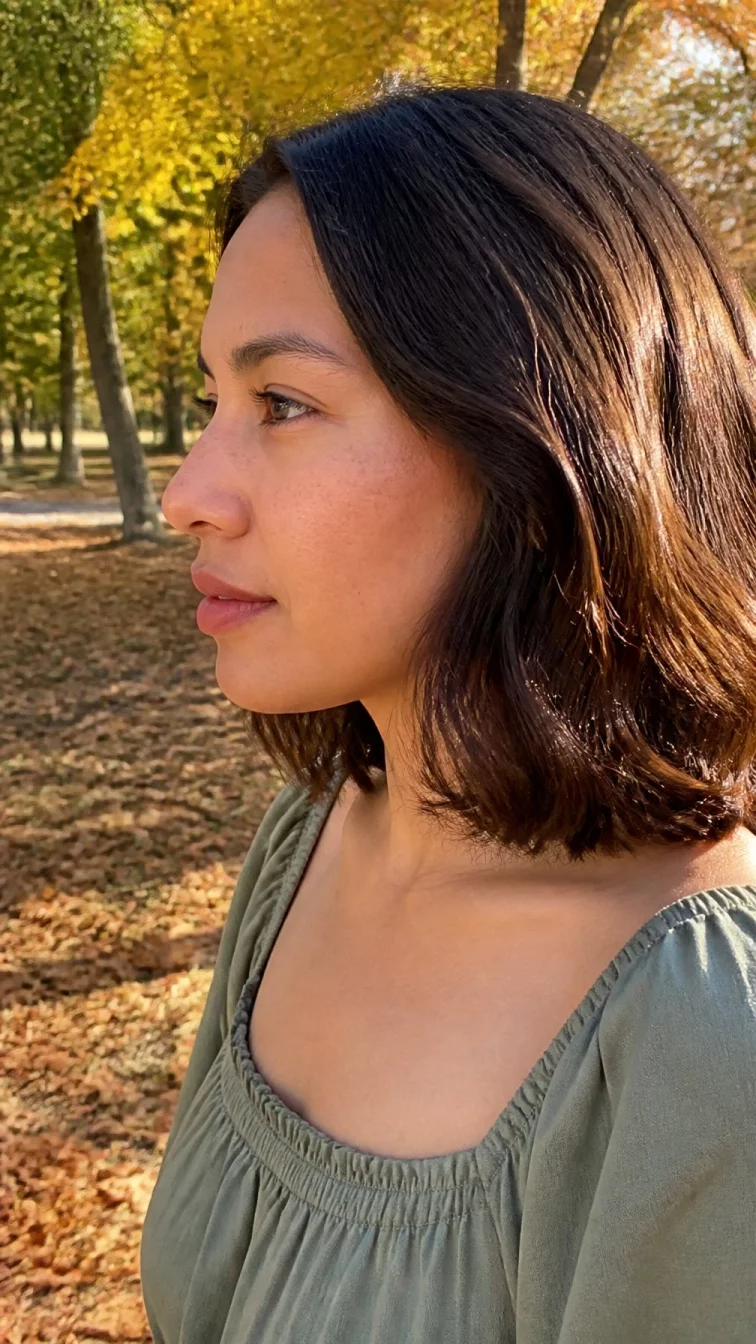 A side photo of a 37-year-old Latina woman with a pear-shaped face, light, narrow nose, straight eyebrows, and wide lips. Her face features faint freckles across the nose, subtle acne marks, and subtle natural facial asymmetry. She has monolid hazel eyes, pronounced cheekbones, neutral expression, cheerful. She is slim and is wearing a sage green square-neck blouse with smoked walnut brown collarbone-length wavy hair, bright natural daylight, outdoor fall background