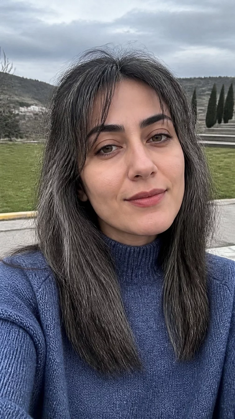 selfie of a 33-year-old Middle Eastern woman with a diamond face, fair, broad nose, thick natural eyebrows, and thin lips. Her face features uneven eyebrows, subtle acne marks, and subtle natural facial asymmetry. She has deep-set hazel eyes, rounded cheeks, subtle smile, thoughtful. She is slim and is wearing a denim blue mock neck sweater with charcoal and shimmering pewter antique silver fox blunt fringe hair, overcast daylight, outdoor