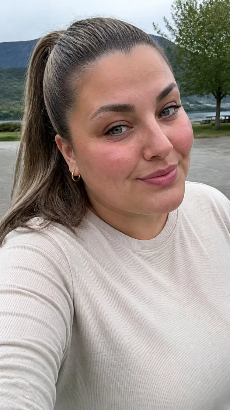 selfie of a 33-year-old Southern European woman with a round face, golden brown, broad nose, thin arched eyebrows, and heart-shaped lips. Her face features small scar on the eyebrow, slightly flushed cheeks, and subtle natural facial asymmetry. She has downturned gray eyes, pronounced cheekbones, closed-mouth smile, playful. She is plus-size and is wearing a cream long sleeve t-shirt with ash brown frosted walnut brown sleek ponytail hair, overcast daylight, outdoor
