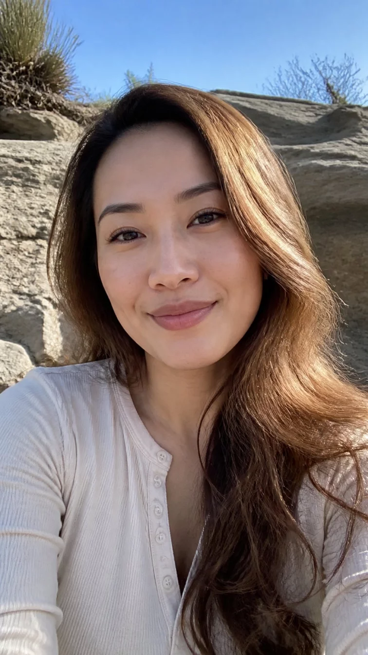 selfie of a 34-year-old East Asian woman with a oblong face, light, straight nose, feathered eyebrows, and wide lips. Her face features dimples, visible pores, and subtle natural facial asymmetry. She has almond-shaped dark brown eyes, soft cheekbones, subtle smile, relaxed. She is average and is wearing a cream henley shirt with bronzed butterscotch wavy hair, bright natural daylight, outdoor